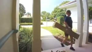 Man wearing sunglasses carrying a long cardboard box on a house porch during daytime