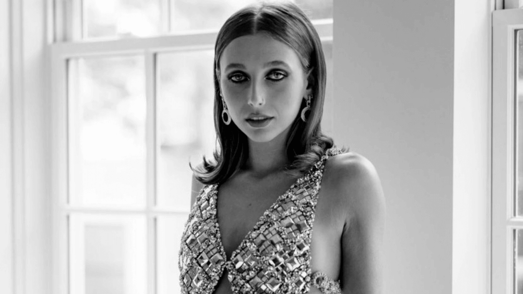 Young woman with sleek hair wearing a beaded, reflective dress and hoop earrings standing by a window