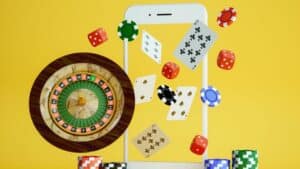Smartphone with floating casino elements including roulette wheel, poker chips, dice, and playing cards on yellow background
