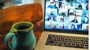 Green ceramic mug filled with coffee next to a laptop showing a virtual meeting with multiple participants.