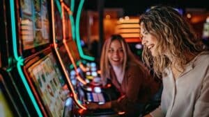 Two women smiling and playing slot machines in a casino with colorful neon lights.