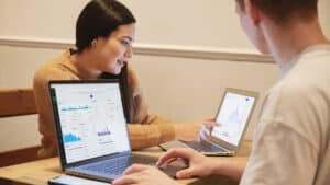 Two people analyzing financial charts on laptops at a wooden table, one pointing at a graph on the screen.