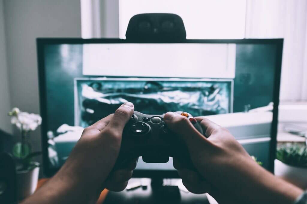 Hands holding a black game controller playing a video game on a monitor in a home setting