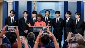 BTS members in black suits with a woman in an orange dress speaking at the White House podium in Washington