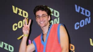 Smiling man wearing glasses and a colorful sleeveless sweater making a peace sign at VidCon event.