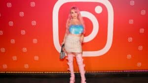 Woman with pink hair wearing a blue sequin top, sparkly skirt, and pink thigh-high boots posing in front of Instagram logo backdrop