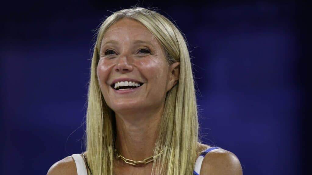 Gwyneth Paltrow smiling with long blonde hair and a gold chain necklace against a dark blue background