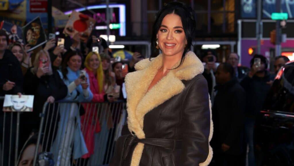 Katy Perry smiling and wearing a brown shearling coat while fans take photos behind barricades at an event.