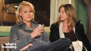 Two women sitting and talking, one holding a glass, both wearing winter coats indoors