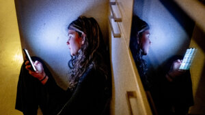 Young woman sitting on the floor using a smartphone, reflected in a nearby mirror or glass surface.