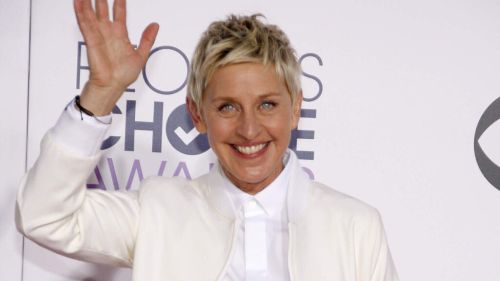 Ellen DeGeneres smiling and waving at the People's Choice Awards wearing a white jacket and shirt.