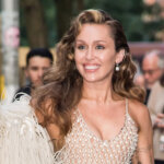 Miley Cyrus smiling and waving, wearing a white fringed outfit with a crowd in the background.