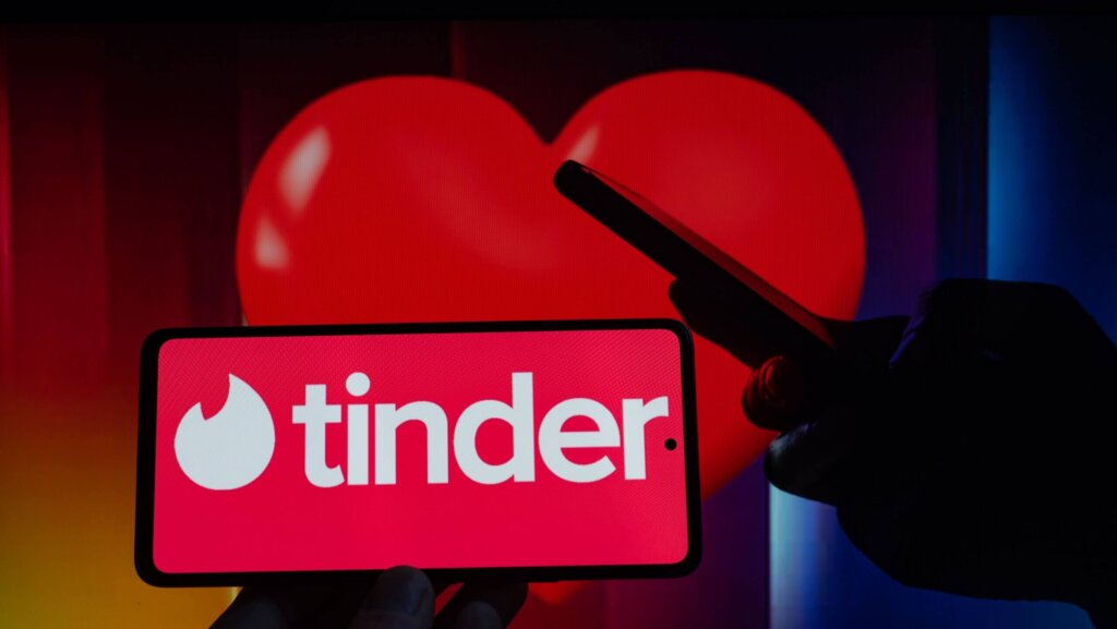 Smartphone displaying Tinder logo with a large red heart in the background and a person holding another phone.