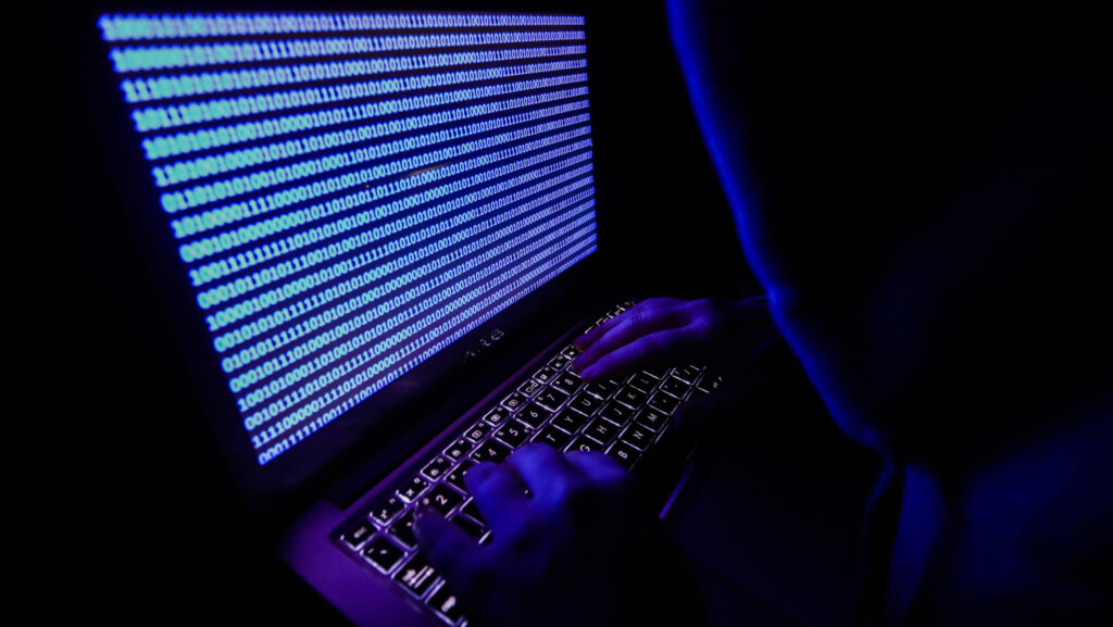 Person in a dark hoodie typing on a backlit laptop keyboard displaying blue binary code on the screen