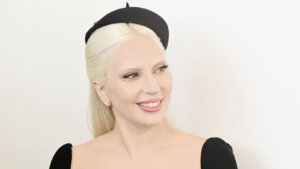 Lady Gaga smiling wearing a black dress and a small black hat against a white background