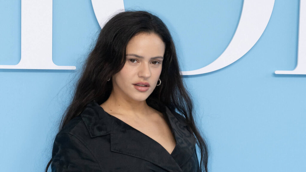Rosalía wearing a black outfit posing against a blue background with white letters.