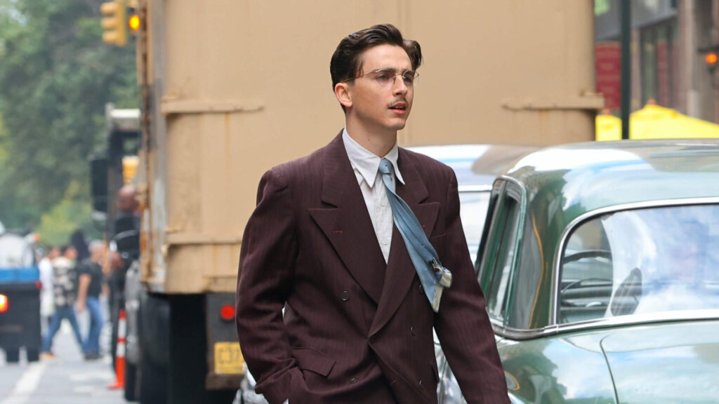 Timothée Chalamet walking on a city street wearing a brown suit and blue tie.