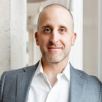 Smiling bald man in a gray suit jacket and white shirt standing by a window and concrete wall