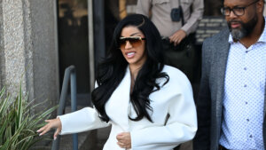 Cardi B wearing a white jacket and large sunglasses walking beside a man in a gray coat and patterned shirt outdoors