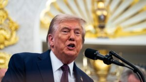 Donald Trump speaking at a microphone with an ornate gold and white background.