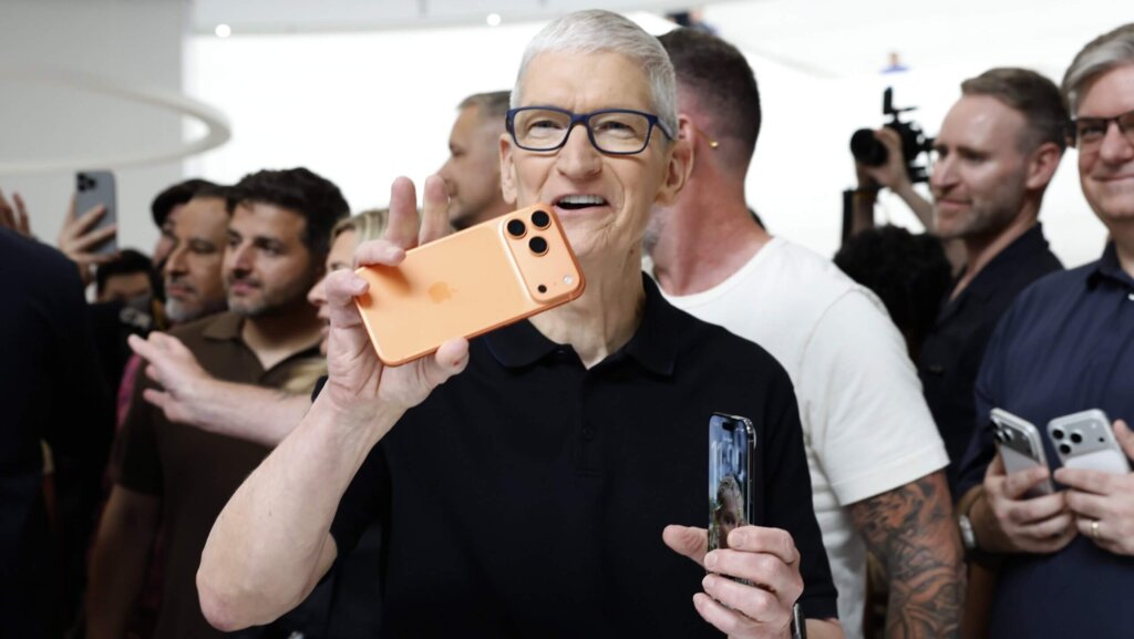 Tim Cook holding an orange iPhone 15 Pro Max at an Apple event with people and photographers in the background.