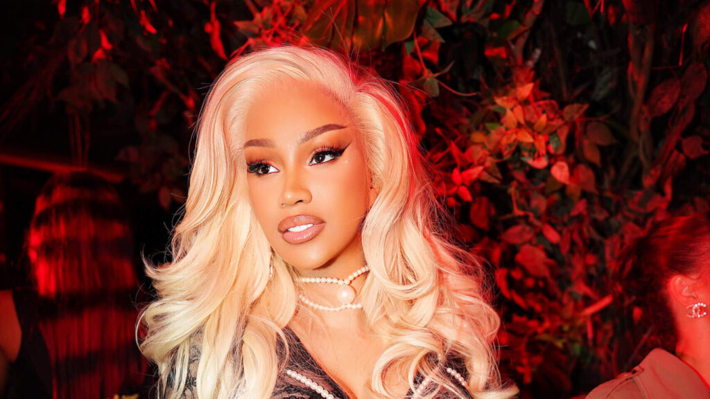 Woman with long platinum blonde hair and pearl necklace in red-lit setting with foliage background