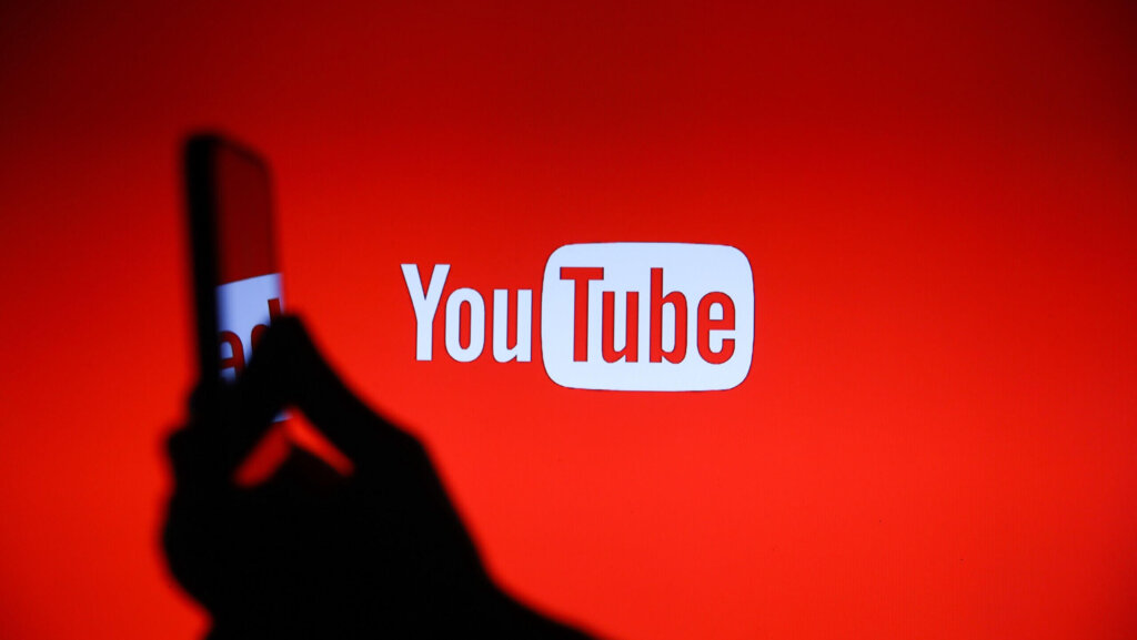 Silhouette of a hand holding a smartphone in front of a red screen displaying the YouTube logo.