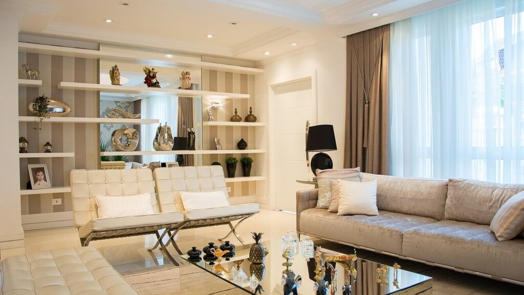 Modern living room with beige sofa, white chairs, glass coffee table, and decorative shelves with figurines and plants.