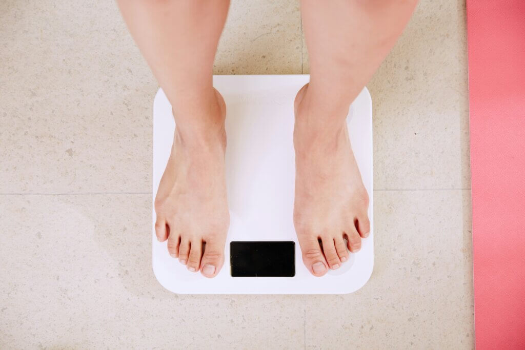 Person standing barefoot on a white digital bathroom scale on a tiled floor next to a pink mat.