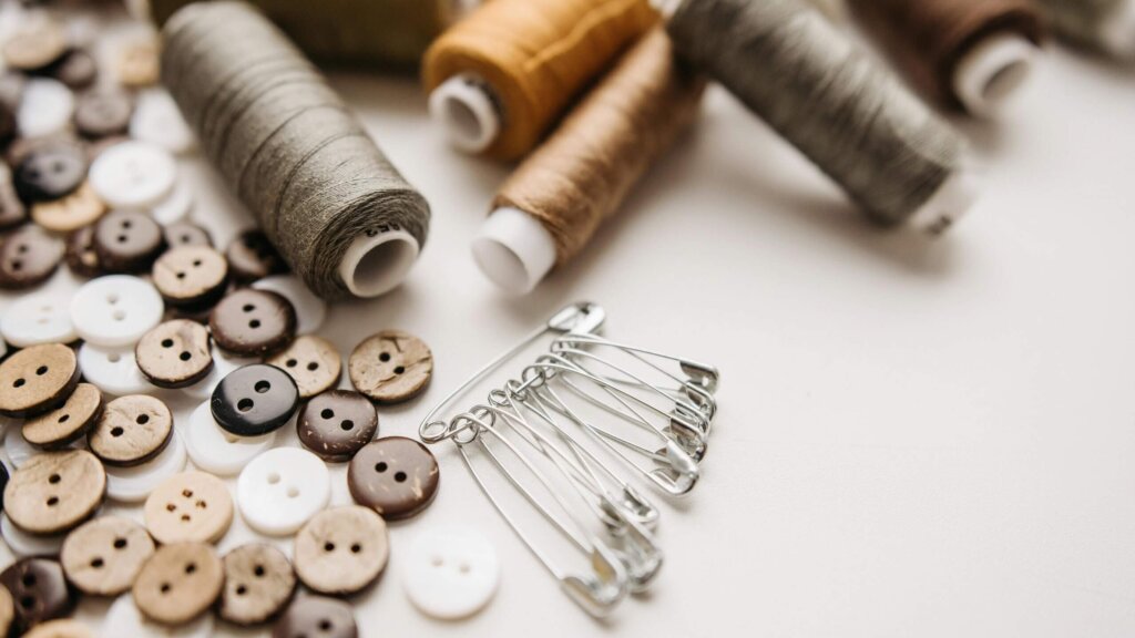 Assorted brown, white, and black buttons, silver safety pins, and spools of thread in neutral tones on a white surface.