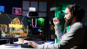 Bearded man wearing a gaming headset celebrating a win on a computer game with "WINNER" displayed on the monitor.