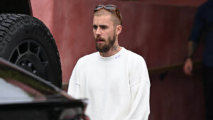 Man with beard and sunglasses on head wearing white sweater and pants standing near black Lamborghini car