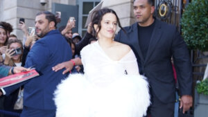 Rosalía in a white feathered dress walking with security and fans taking photos around her