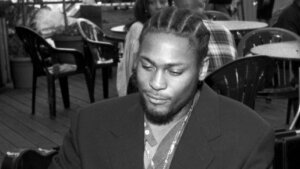 Man with braided hair wearing a suit jacket, holding a cigarette and lighter, seated outdoors at a cafe.