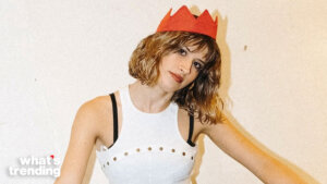 Woman with wavy hair wearing a red paper crown and a white sleeveless top with black straps against a plain background