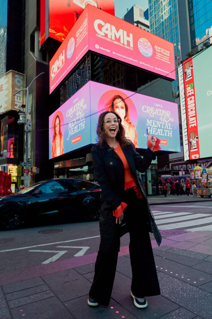 Woman smiling and pointing at a Times Square billboard promoting Creators 4 Mental Health campaign.