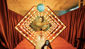 Person taking photo of another person standing in illuminated Spotify-themed art installation with red curtains.