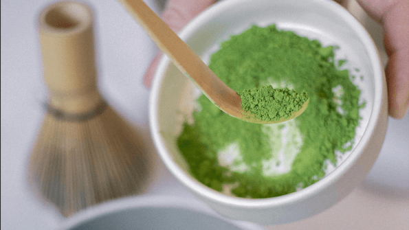 Close-up of a bamboo scoop holding green matcha powder above a white bowl with a bamboo whisk in the background.