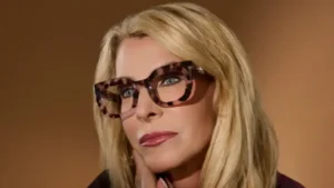 Blonde woman with tortoiseshell glasses resting her chin on her hand against a brown background