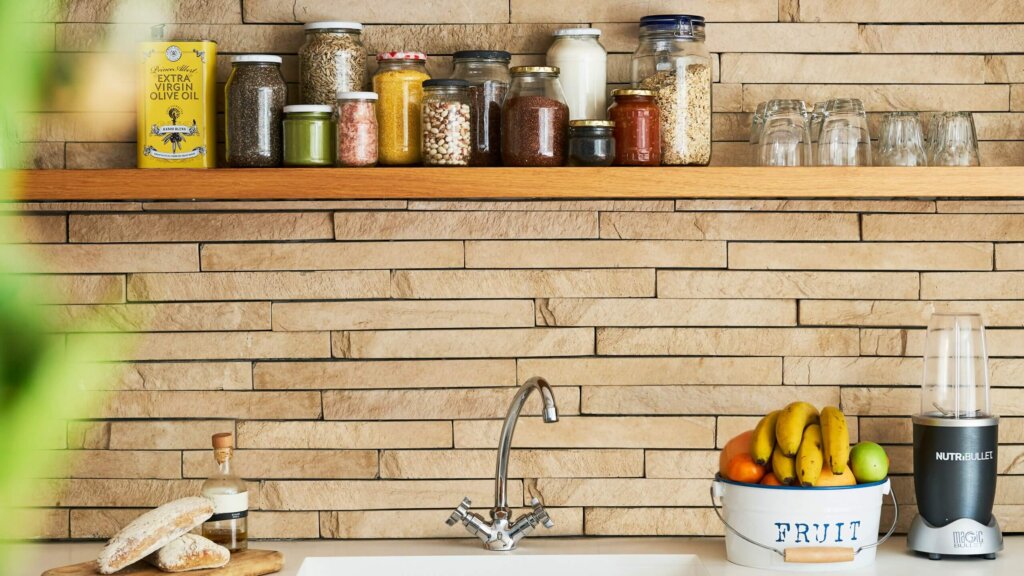 Kitchen countertop with NutriBullet blender, fruit bucket, bread, olive oil, and jars of spices and grains on a wooden shelf