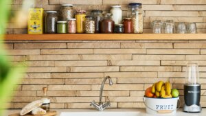 Kitchen countertop with NutriBullet blender, fruit bucket, bread, olive oil, and jars of spices and grains on a wooden shelf