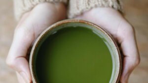 Hands holding a ceramic cup filled with green matcha tea against a blurred background.