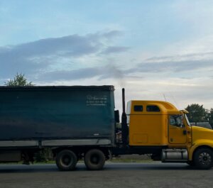Yellow semi-truck with a blue trailer emitting black smoke on a rural road at dusk