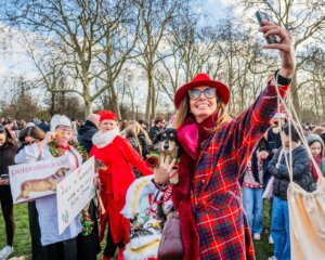 Woman in red plaid coat and hat taking a selfie while holding a dog at a festive outdoor gathering with people in holiday attire.