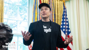 Elon Musk speaking with hands raised in a room with American flags and yellow curtains behind him