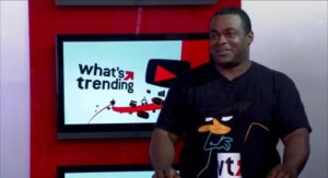 Man wearing a Perry the Platypus t-shirt standing next to a screen displaying "what's trending" logo
