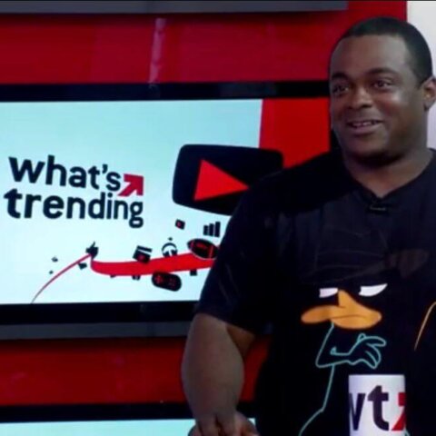 Man wearing a Perry the Platypus t-shirt standing next to a screen displaying "what's trending" logo